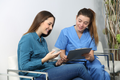 nurse and female patient looking at clipboard as they sit in chairs together nurse and female patient looking at clipboard as they sit in chairs together