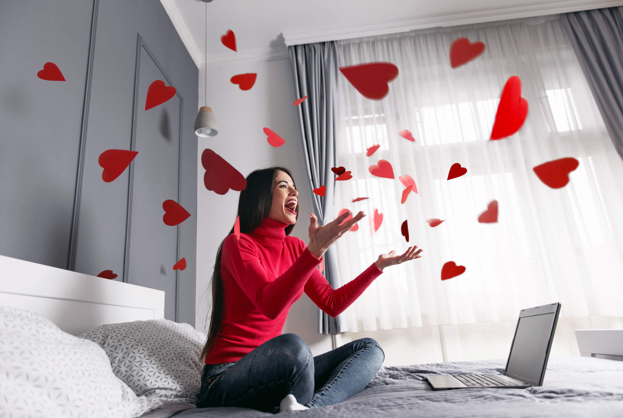 black and white image of woman on bed with laptop tossing red hearts in air black and white image of woman on bed with laptop tossing red hearts in air