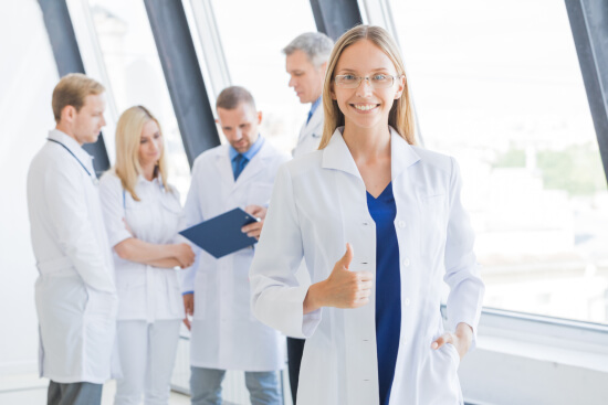 woman in lab coat giving thumbs up with a happy group of healthcare professionals behind her woman in lab coat giving thumbs up with a happy group of healthcare professionals behind her