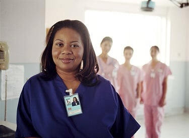 Confident nurse in blue scrubs in foreground; nurses in pink scrubs in background Confident nurse in blue scrubs in foreground; nurses in pink scrubs in background