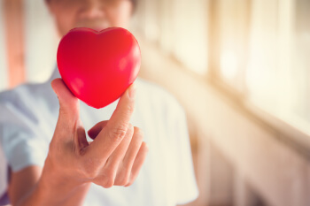 heart held in fingers in foreground; woman in scrub in background heart held in fingers in foreground; woman in scrub in background