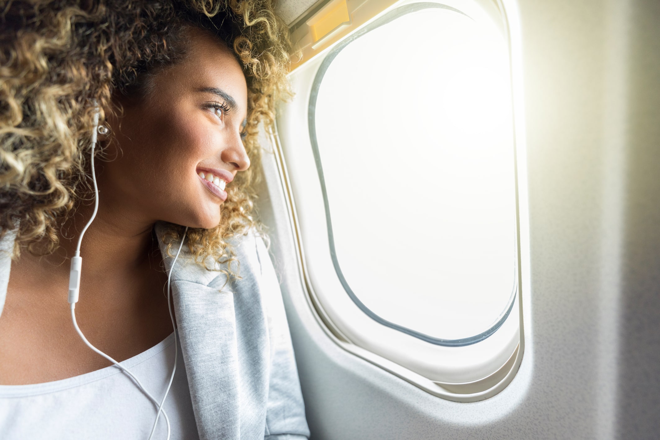 beautiful travel nurse female with curly hair stares out the airplane windows with an optimistic smile on her face
